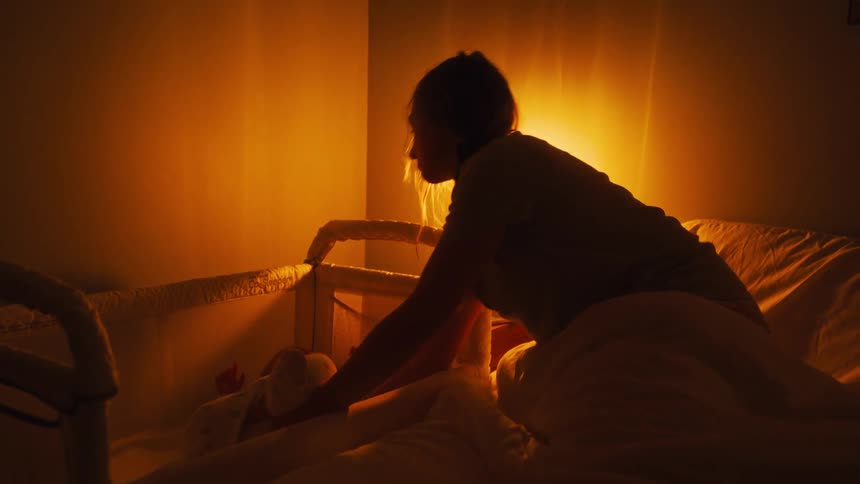 A mother lights an oil lamp in the middle of the night to pick up her crying baby from its bassinet next to her bed