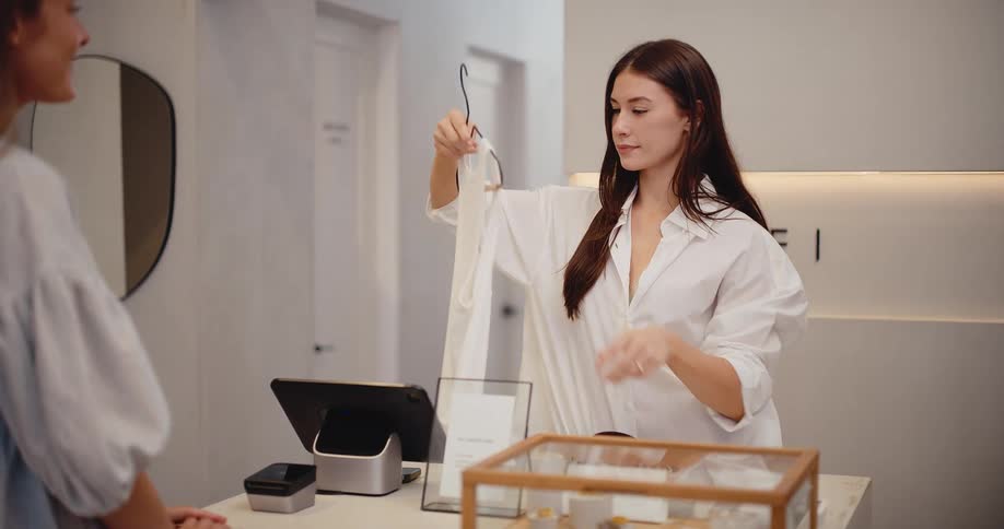 Slow motion of a friendly sales assistant taking a white top and belt from a customer to process a purchase in a modern boutique
