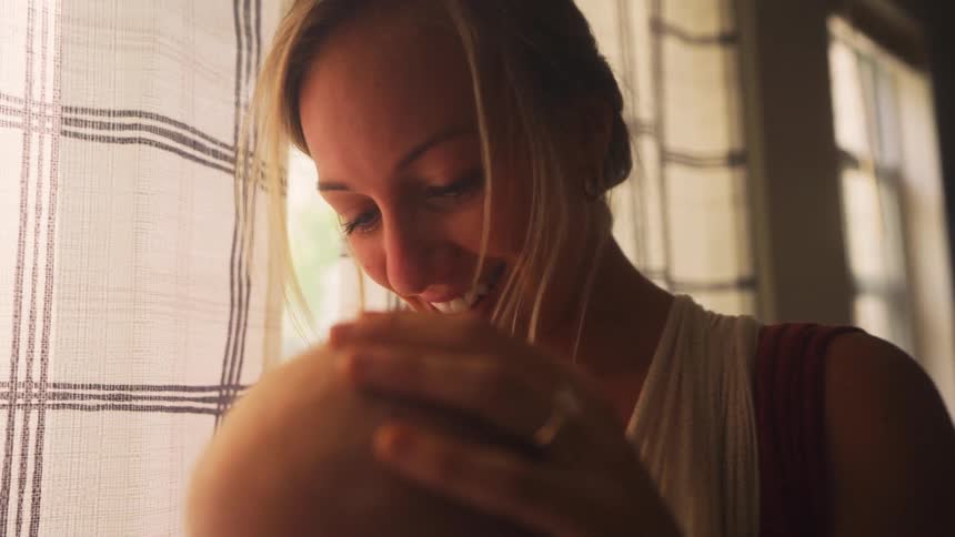 A young mother smiles lovingly at her newborn baby, gently caressing their head in a tender, sunlit moment by a window