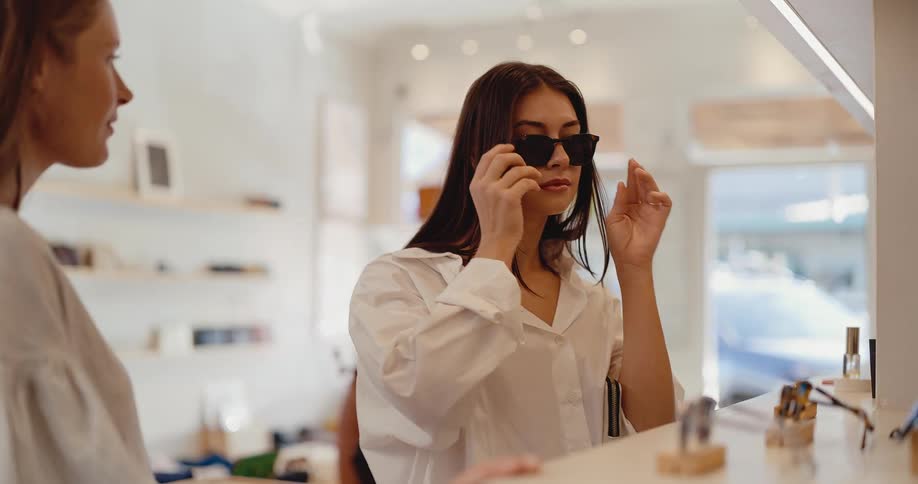 A stylish young woman tries on a pair of sunglasses in a modern boutique and smiles happily while her friend watches