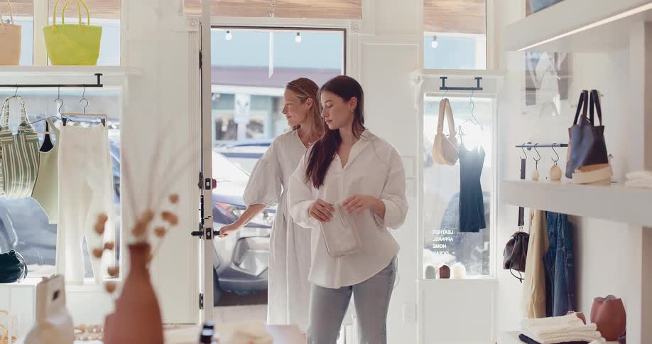 Two young women walk through the door of a bright and airy clothing boutique in slow motion