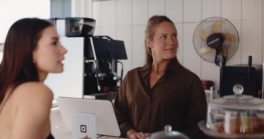 A friendly female barista smiles while taking an order from a customer at the counter of a bright and modern coffee shop