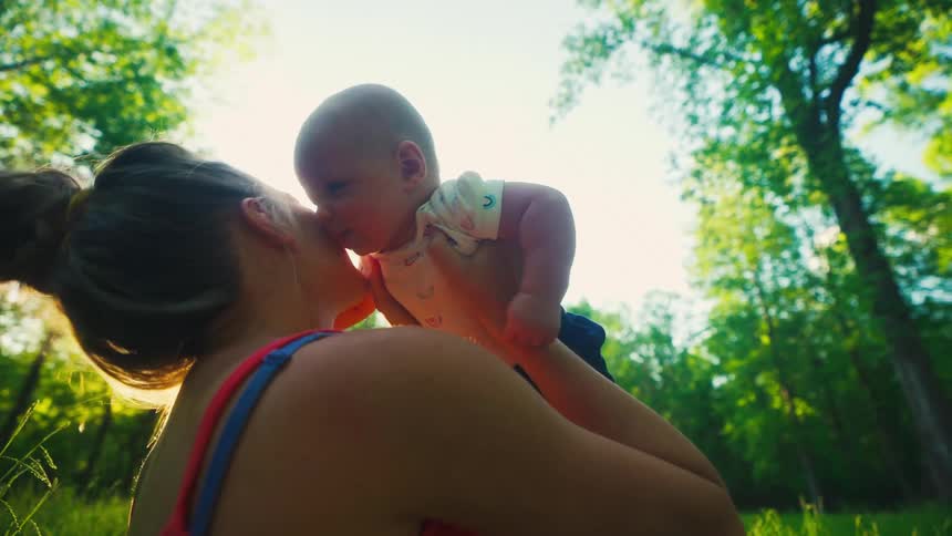 A loving mother lifts her newborn baby up into the air and kisses them in a sunlit forest in slow motion