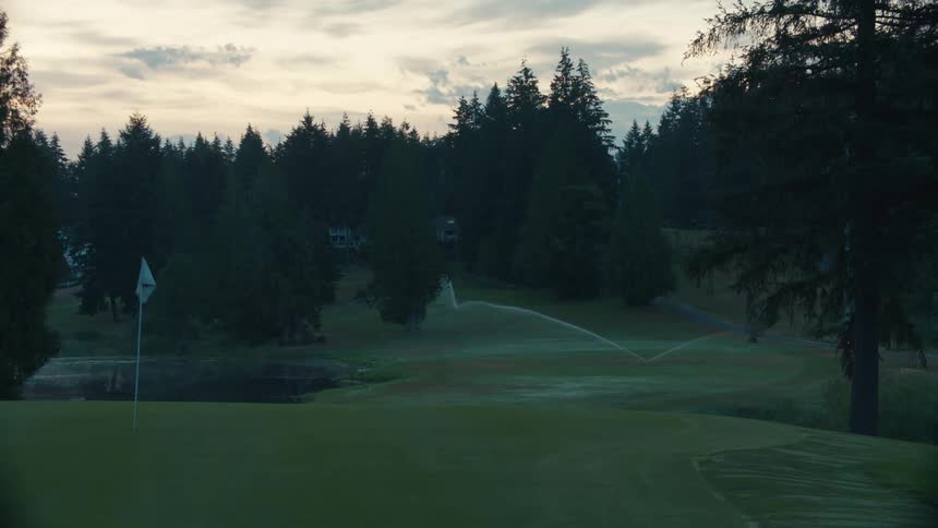 Early morning sprinklers water a tranquil golf course with a putting green and a lush forest in the background