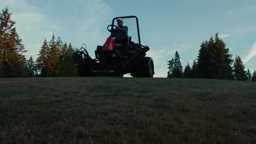 A groundskeeper drives a large ride-on mower over a grassy hill towards the camera at dawn