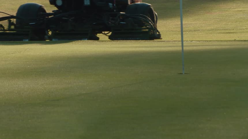 A groundskeeper uses a professional ride-on lawn mower to cut the grass on a putting green at a golf course