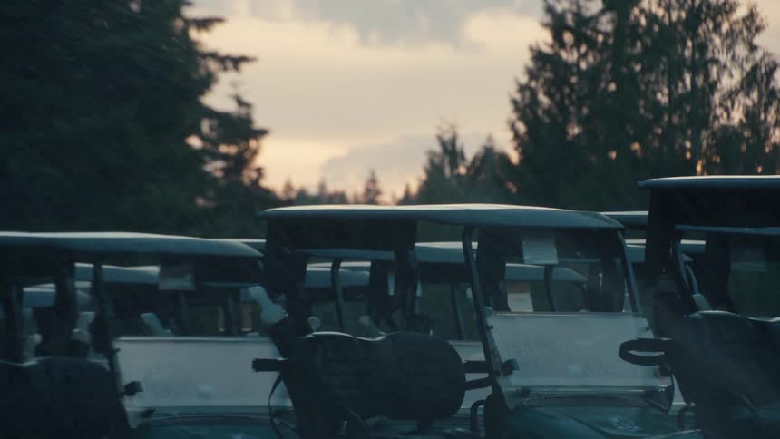A line of empty golf carts sits idle on a course during a rain shower at sunset