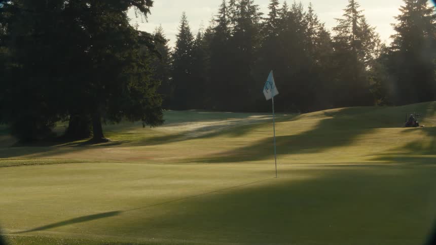 A serene golf course green in the early morning sunlight, with a groundskeeper mowing the fairway in the background