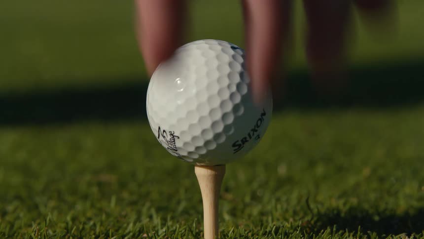 A golfer's hand places a wooden tee in the grass and sets a golf ball on top in slow motion