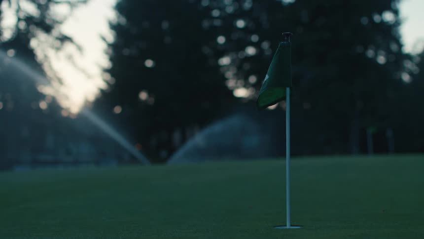 A golf ball sinks a putt on a serene green at dusk while sprinklers water the course