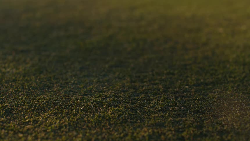 A golf putter strikes a ball on a lush green in dramatic slow motion during sunset