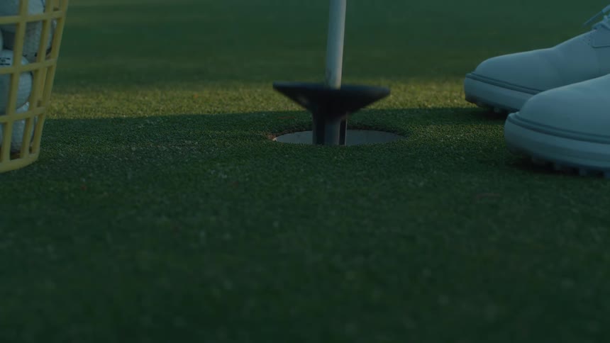 A golfer retrieves a cup full of practice balls from a hole on the putting green in slow motion