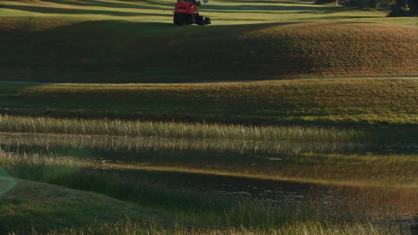 A groundskeeper operates a fairway mower across the rolling green hills of a golf course next to a tranquil pond at sunrise