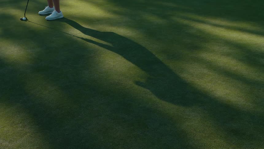The shadow of a golfer stretches across the putting green as they make a shot in slow motion