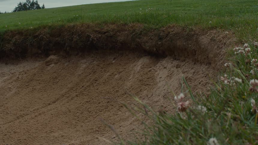 A golf ball rolls into a deep sand bunker in slow motion on a golf course