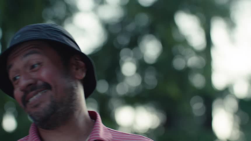 A happy man wearing a bucket hat turns his head and laughs playfully while standing outside in nature