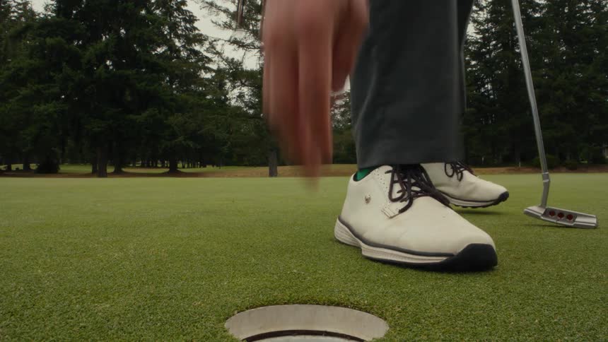 From a low angle, a golfer in slow motion sinks a putt, retrieves the ball from the hole, and replaces the flag
