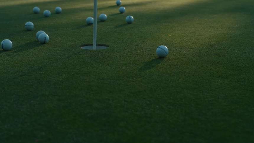 Multiple golf balls roll across a putting green in slow motion, with one successfully sinking into the hole