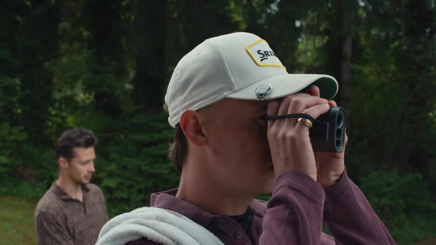 A golfer wearing a white cap uses a rangefinder to scope the distance on the course as his friend looks on
