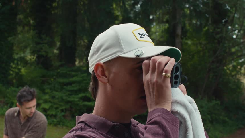 A golfer with a mustache and mullet uses a rangefinder to scope out the course while his friend waits