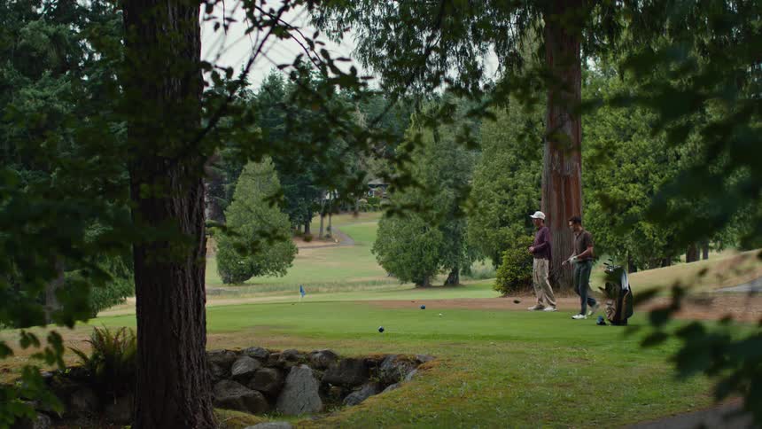 Two men take turns teeing off on a beautiful, lush golf course surrounded by tall trees