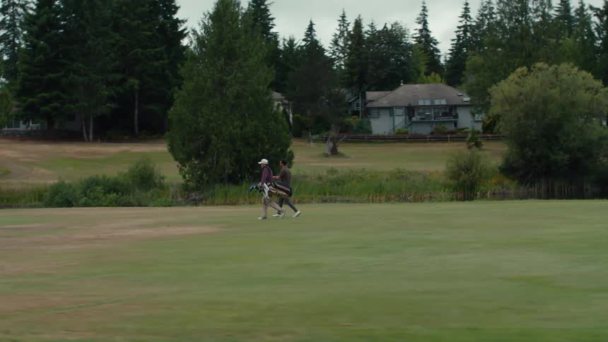 A long tracking shot follows two men walking and talking across a vast, green golf course with their bags