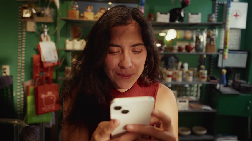 A smiling woman with long dark hair enjoys content on her smartphone while sitting in a cozy, eclectic shop