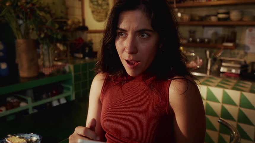 A young woman's face lights up with surprised excitement as she receives good news on her smartphone while sitting in a cozy, eclectic cafe