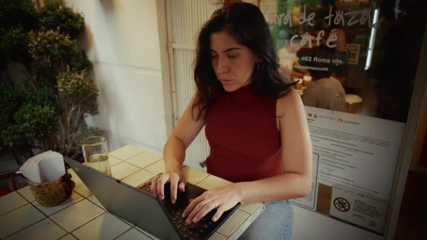 A young woman sits down at an outdoor cafe table, opens her laptop, and begins to work intently