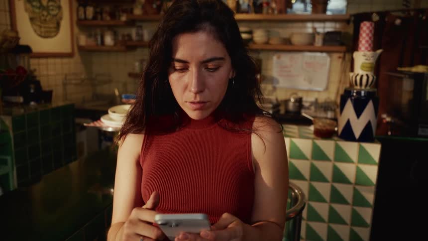 Slow zoom on a concerned woman sitting at a counter in a retro kitchen while intently scrolling on her smartphone
