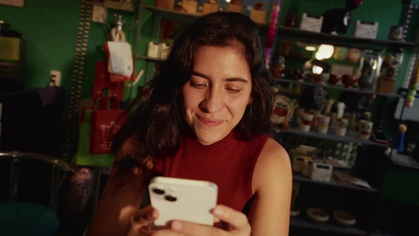 A woman's expression softens into a happy smile as she scrolls on her smartphone inside a cozy, sunlit shop