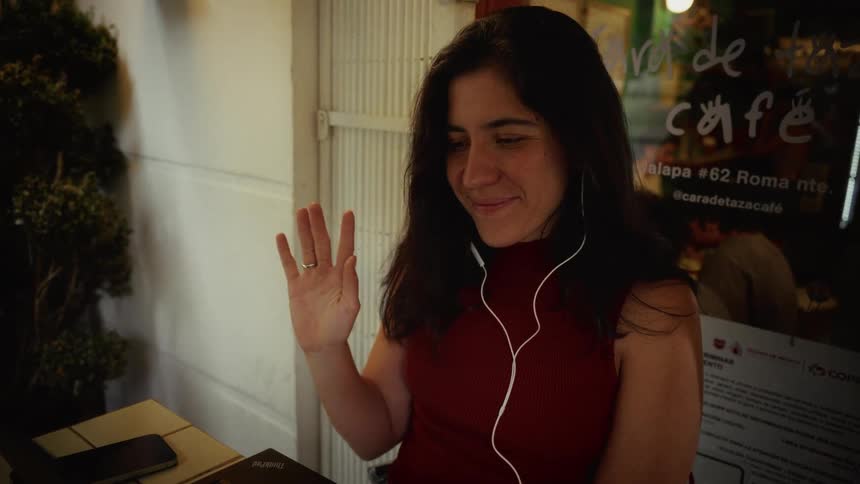 A woman smiles and waves while on a video call on her laptop at an outdoor cafe at night