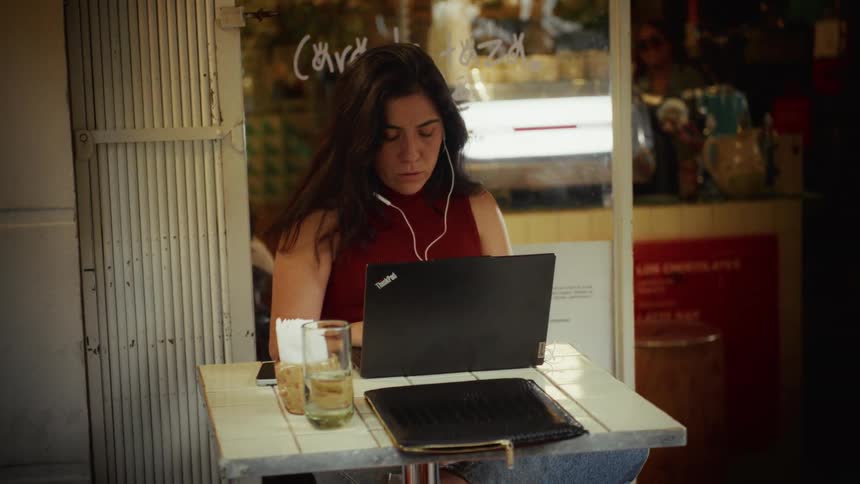 A young woman wearing earphones works intently on her laptop at an outdoor cafe table