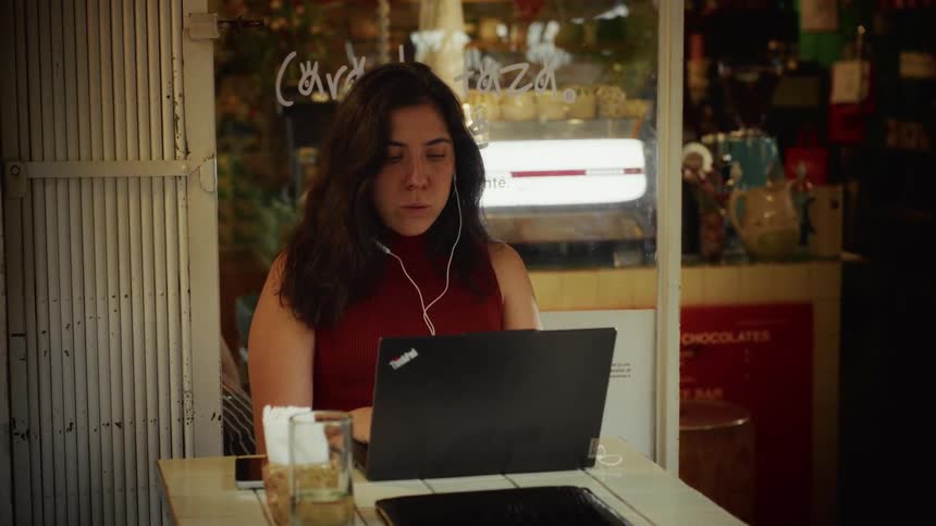 A woman with dark hair works on her laptop with headphones in at an outdoor cafe table