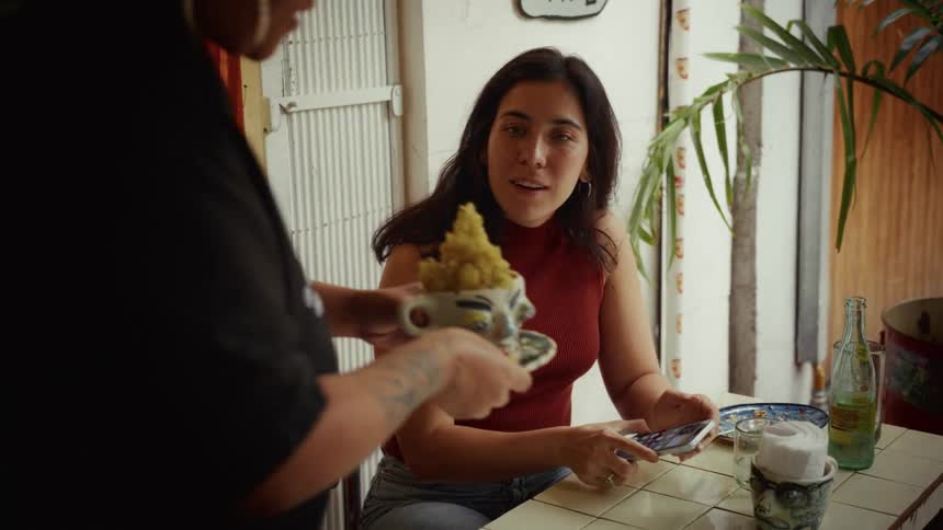 A woman at a cafe looks up from her phone in surprise and delight as a server presents her with an artistic dish