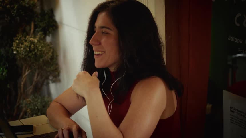 A woman intently watching her laptop in a cafe breaks into a smile while listening with earphones