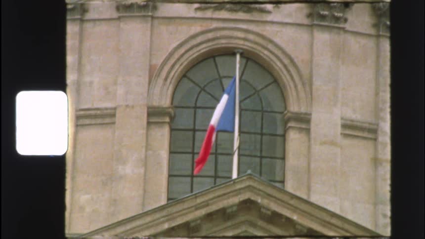 A vintage film shot captures the French flag waving gently in front of an arched window on a historic building