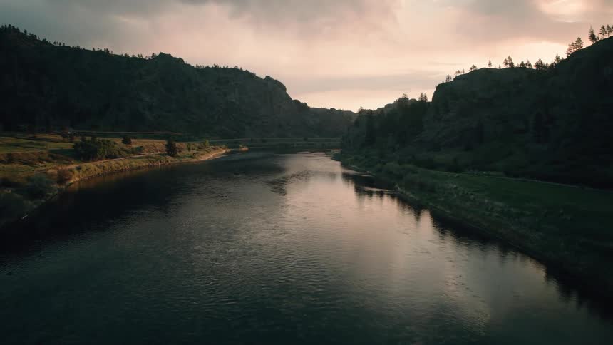 An atmospheric drone shot slowly flies over a truss highway bridge down a tranquil river canyon at dawn