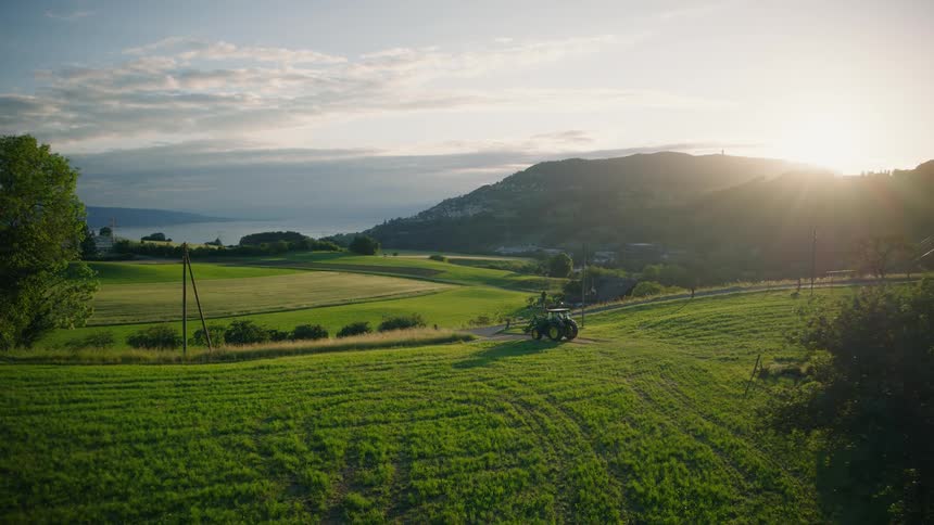 A serene drone shot captures a tractor driving through a lush green valley at sunset, with rolling hills and a distant lake in the background