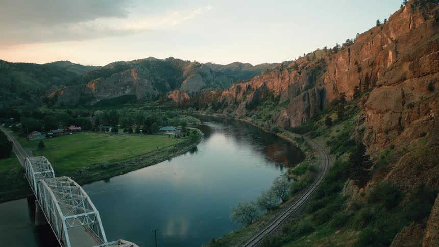An aerial view slowly moving towards a steel bridge crossing a wide river nestled in a rocky mountain canyon at sunset