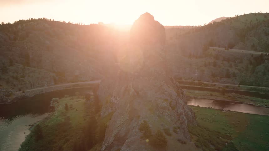 A majestic aerial view shows the sun setting behind a towering rock formation next to a winding river in a mountain valley