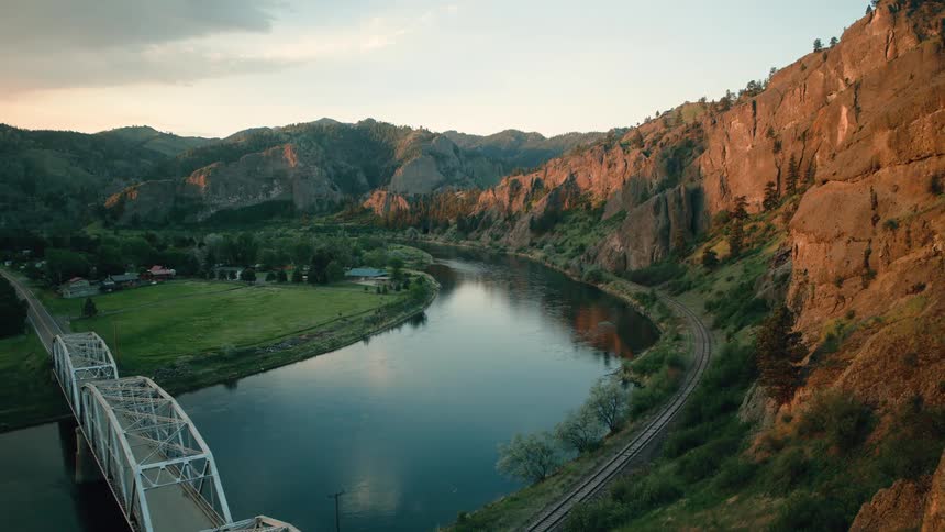 An aerial view pulls back over a winding river in a mountain valley to reveal a steel truss bridge at golden hour