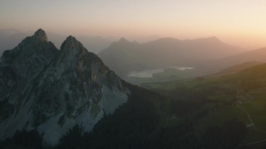 An epic aerial view pulls back from a rugged mountain peak to reveal a vast, serene valley and a distant lake glowing in the warm light of sunset