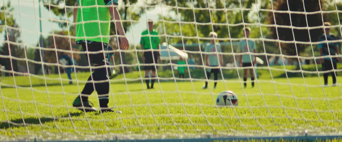 From behind the goal net, a young boy takes a penalty kick during a youth soccer match