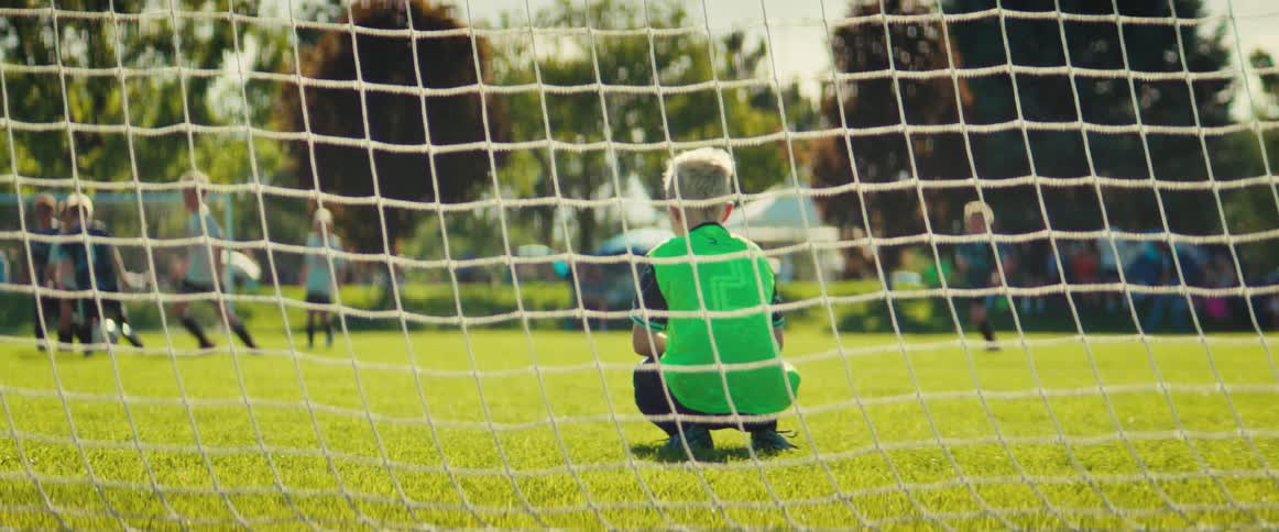 View from behind the goal as a young boy in a bright green jersey plays goalie during a kids' soccer match