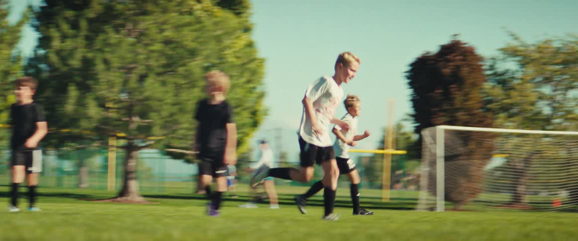 A dynamic tracking shot follows a team of young boys as they race down the soccer field on a sunny day to score a goal
