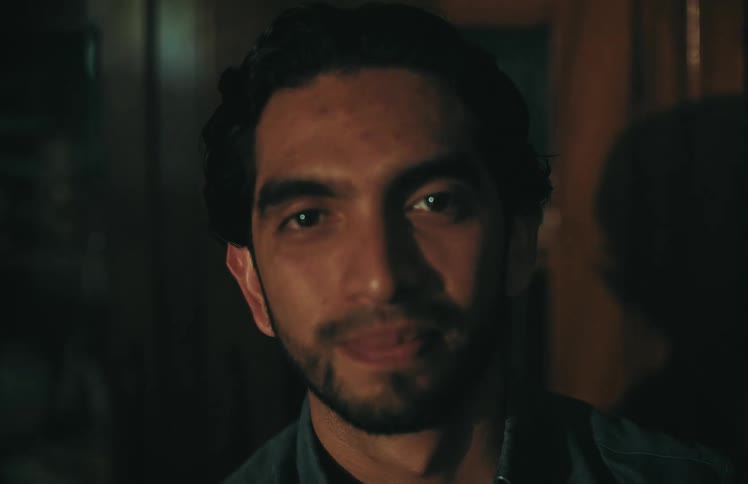 A man with a beard looks directly into the camera as a single light source flickers across his face in a dark room