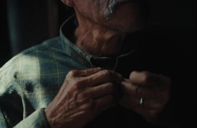 An elderly man with weathered hands and a white mustache slowly buttons his plaid shirt in a dramatically lit room