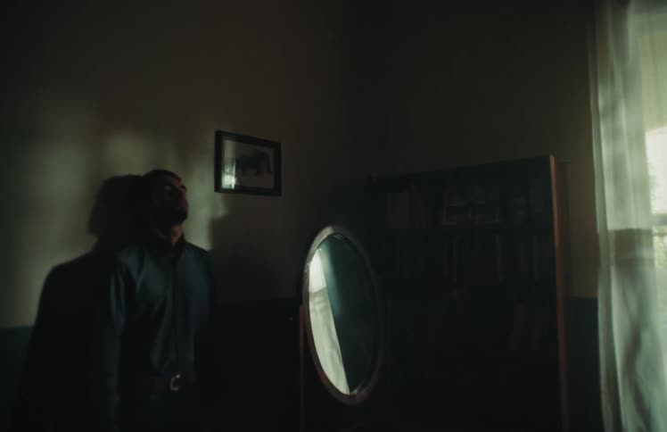 A pensive man stands alone in a shadowy room, his silhouette cast on the wall by a sliver of sunlight from the window