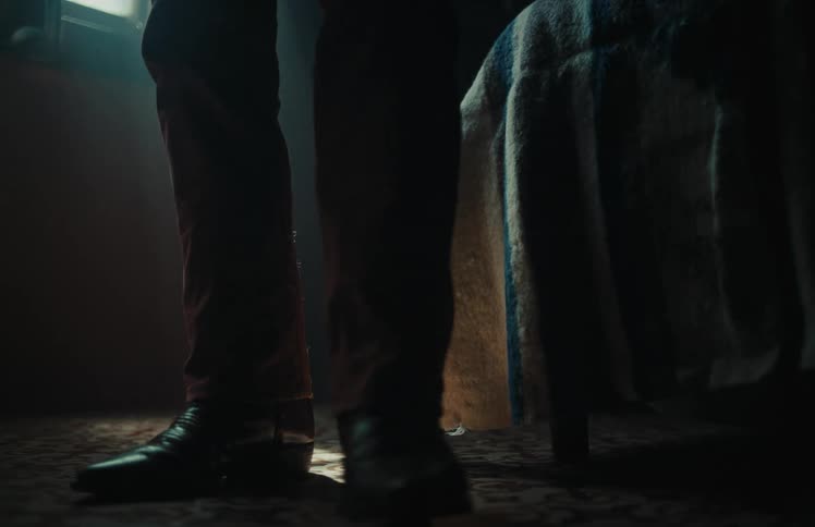 A person stands up from a chair and walks towards the camera in a dark, dusty room illuminated by a single sunbeam